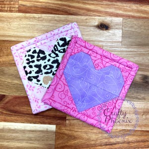 May include: Two pink fabric coasters with a heart-shaped design. One coaster features a purple heart with a swirl pattern, and the other has a black and white leopard print heart. The coasters have a pink border with a white swirl pattern.