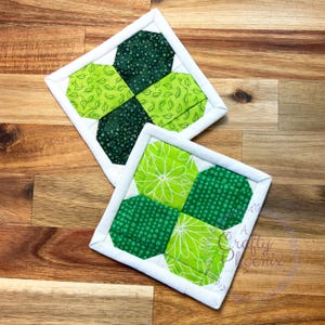 May include: Two green and white quilted pot holders with a four-leaf clover design. The pot holders are on a wooden surface.