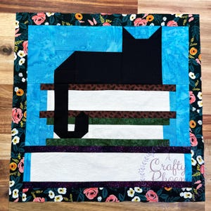 Puede incluir: Un edredón cuadrado con un diseño de gato negro sobre una pila de libros, sobre un fondo azul. El edredón está enmarcado por un borde floral en rosa, blanco y amarillo sobre negro. El texto "A Crafty Phoenix" es visible en el edredón.