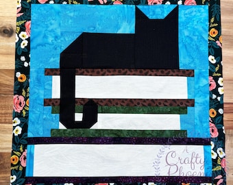 May include: A square quilt featuring a black cat silhouette atop a stack of books. The background is blue, and the quilt is bordered with a floral pattern. The text "A Crafty Phoenix" is visible in the lower right corner.