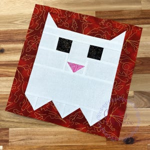 May include: A square quilt block featuring a stylized white cat face with black speckled eyes and a pink patterned nose. The cat is framed by a deep red border decorated with a gold-colored outline of oak leaves and swirling vine patterns.