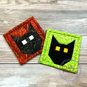 May include: Two black cat-shaped quilted coasters. One coaster has a red background with gold leaf accents and the other has a green background with a leaf pattern. Both coasters have white eyes and yellow eyes.