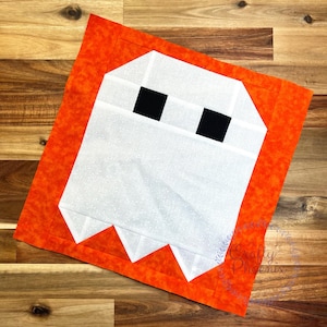 Könnte beinhalten: Ein quadratischer Stoff-Quiltblock mit einer weißen Geisterfigur, zwei schwarzen quadratischen Augen und spitzen weißen Zähnen am unteren Rand. Der Geist ist auf einem leuchtend orangefarbenen Hintergrund mit gesprenkelter Textur zentriert.