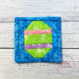 Pode incluir: Um descanso de copo acolchoado azul e verde com um padrão floral. O descanso de copo tem a forma de um ovo e tem uma faixa rosa e roxa.