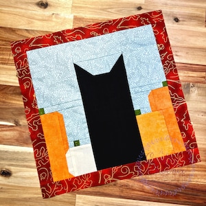 May include: A quilted square featuring a black cat silhouette, orange pumpkins, and a light blue background with a floral pattern. The quilt has a red border with a gold leaf design, perfect for fall decor.