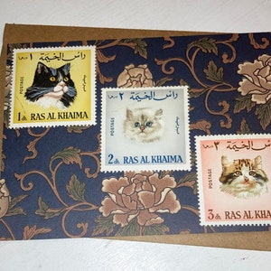 Puede incluir: Tres sellos postales antiguos de Ras Al Khaimah, Emiratos Árabes Unidos, con diferentes razas de gatos. Los sellos están sobre un fondo floral azul y marrón.