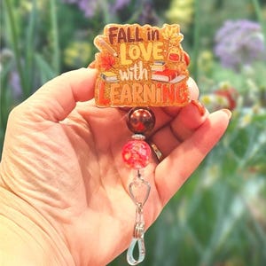 May include: A badge reel with the text "FALL in LOVE with LEARNING". The badge has a fall theme with books and leaves. The reel has two beads, one brown and one red, and a clear clip.