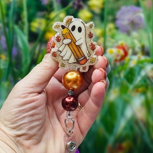 May include: A badge reel featuring a cartoon ghost holding a pencil, surrounded by autumn leaves. The badge reel has a gold and burgundy bead accent. The design is on a clear, glittery background.