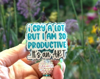 May include: A square badge reel featuring a teal glitter background and white text that reads I CRY A LOT BUT I AM SO PRODUCTIVE ...It's an ART. The badge is attached to a retractable cord decorated with two spherical beads with a textured finish.