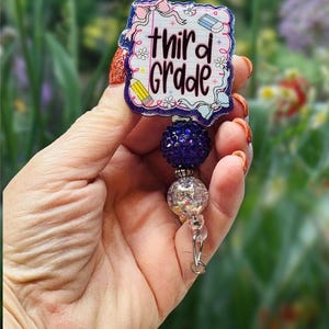 May include: A badge reel with the words "third grade" in a whimsical font. The badge has a white background with a blue border and features school-themed illustrations. The reel has a dark blue beaded accent and a clear bead with a silver clasp.