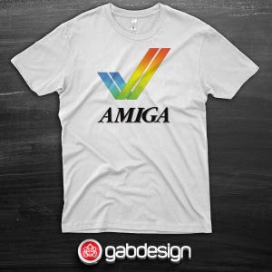 Può includere: Una t-shirt bianca a maniche corte con la grafica di un segno di spunta stilizzato con un gradiente arcobaleno blu, verde, giallo e arancione. Sotto il segno di spunta, la parola AMIGA è stampata in un carattere serif nero e grassetto.