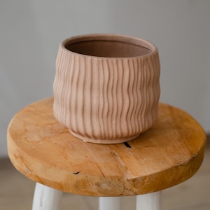 Può includere: Un vaso di terracotta rosa chiaro con una texture ondulata e nervata. Il vaso è appoggiato su uno sgabello di legno.