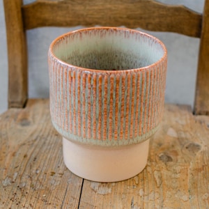 Op de afbeelding: Een keramische plantenpot met een lichtgroene en oranje glazuur. De pot heeft een geribbeld ontwerp en een brede basis.