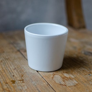May include: A white ceramic cup with a smooth, rounded rim. The cup is sitting on a wooden surface.
