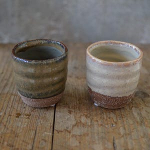 Può includere: Due tazze in ceramica fatte a mano dall'aspetto rustico. Una è marrone e verde, l'altra beige e marrone. Entrambe hanno un design strutturato e stratificato, poste su una superficie di legno.