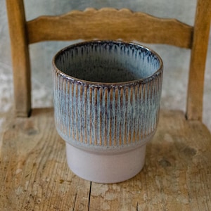 Può includere: Un vaso in ceramica con un design a righe verticali blu chiaro e marrone. Il vaso ha una base rosa chiaro e un bordo blu scuro. È appoggiato su una sedia di legno.
