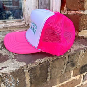 K&ouml;nnte beinhalten: Rosa-wei&szlig;e Trucker-Kappe mit wei&szlig;em Netzr&uuml;cken. Die Kappe hat den Text "What's That" in Gr&uuml;n auf der Vorderseite gestickt.