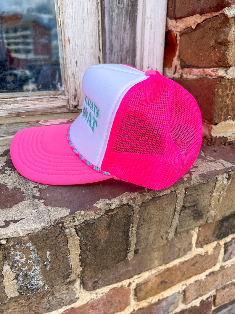 K&ouml;nnte beinhalten: Rosa-wei&szlig;e Trucker-Kappe mit wei&szlig;em Netzr&uuml;cken. Die Kappe hat den Text "What's That" in Gr&uuml;n auf der Vorderseite gestickt.