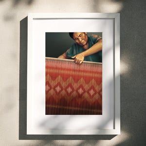 Op de afbeelding: Een ingelijst kunstwerk toont een persoon die een rood en goud textiel weeft met een ruitpatroon. De persoon draagt een groen shirt. Het kunstwerk zit in een witte lijst, gemonteerd op een grijze muur, met schaduwen.