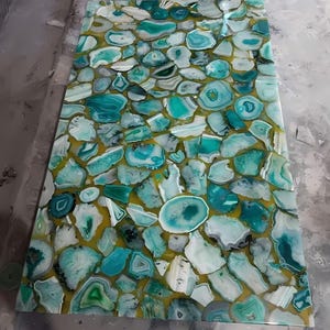 May include: A rectangular tabletop featuring a mosaic of teal, white, and gray agate slices set in a golden-yellow resin. The agate pieces vary in size and shape, creating a unique, decorative surface. The overall design is a blend of natural stone and modern design.