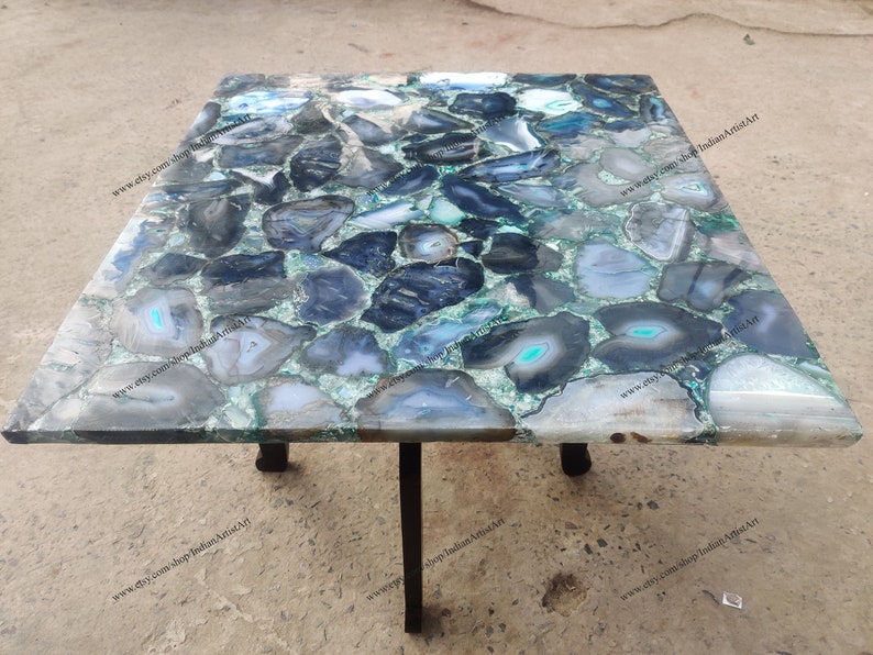 Pu&ograve; includere: Un tavolino da caff&egrave; quadrato con un piano unico in pietra di agata in tonalit&agrave; di blu, verde e grigio. Il tavolo ha una base in metallo nero con quattro gambe.