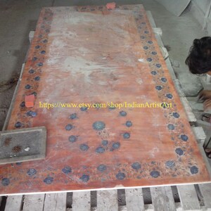 May include: A large, rectangular, red marble table top with a floral design in blue and white. The table top is being worked on by a person in a white shirt and red pants. The text "https://www.etsy.com/shop/IndianArtistArt" is visible on the table top.