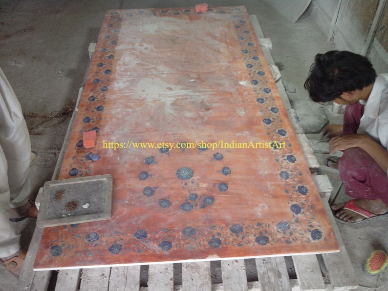 May include: A large, rectangular, red marble table top with a floral design in blue and white. The table top is being worked on by a person in a white shirt and red pants. The text "https://www.etsy.com/shop/IndianArtistArt" is visible on the table top.