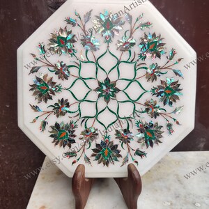 Op de afbeelding: Achtekantige witte marmeren tafelblad met een ingewikkeld bloemmotief ingelegd met parelmoer en groene steen. Het tafelblad staat op een houten standaard.