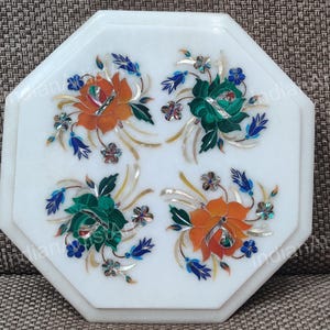 May include: White octagonal marble tabletop inlaid with floral designs. The flowers are orange and green, with blue and silver accents. A decorative piece for home decor.