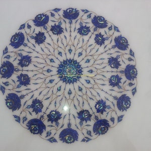May include: A white marble round table top with a blue floral design inlaid with lapis lazuli stones. The design features a central rosette with a repeating pattern of stylized roses.