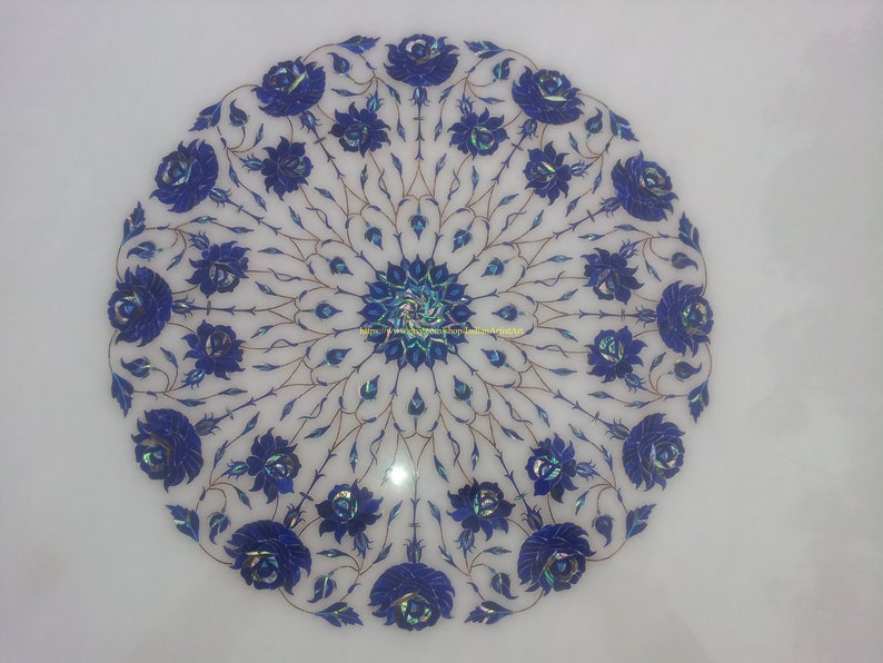 May include: A white marble round table top with a blue floral design inlaid with lapis lazuli stones. The design features a central rosette with a repeating pattern of stylized roses.