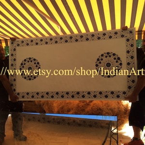 May include: Two people are holding up a large, rectangular, white table top with a blue and white geometric pattern. The table top has a decorative border and two circular designs in the center. The table top is being held up in front of a striped awning.