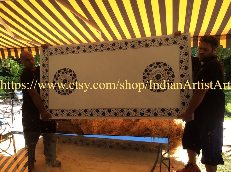 May include: Two people are holding up a large, rectangular, white table top with a blue and white geometric pattern. The table top has a decorative border and two circular designs in the center. The table top is being held up in front of a striped awning.