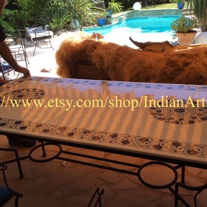 May include: A white marble table with intricate blue and gold floral designs. The table has a black metal frame and is set up outdoors near a swimming pool. The text "https://www.etsy.com/shop/IndianArtistArt" is visible on the table.