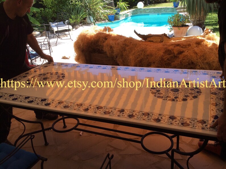 May include: A white marble table with intricate blue and gold floral designs. The table has a black metal frame and is set up outdoors near a swimming pool. The text "https://www.etsy.com/shop/IndianArtistArt" is visible on the table.
