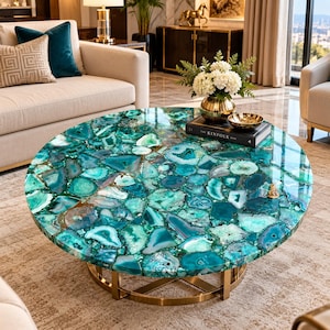 May include: A round coffee table featuring a tabletop composed of polished teal and green agate slices with a glossy finish. The table is supported by a circular gold-toned metal base and is topped with a black book and a gold bowl of white flowers.