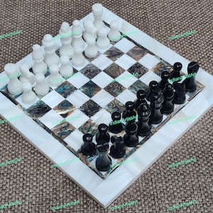May include: A handcrafted chess set with a square board, white border, and a checkerboard pattern. The board has iridescent abalone shell accents. The chess pieces are black and white, ready for a game.
