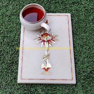 May include: White marble coaster with a red and gold floral design. The coaster is decorated with inlaid mother of pearl. A white teacup with red tea is on top of the coaster.
