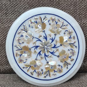 May include: Round white marble coaster with a floral design. The coaster features a detailed inlay of flowers in shades of gold, white, and blue. The design is centered and surrounded by a blue border.