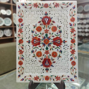 May include: A rectangular white marble decorative tray with intricate floral inlay. The tray features a central floral design and a border of red, orange, blue, and green flowers and leaves. The background has a delicate openwork pattern.