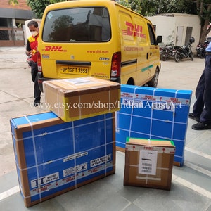 May include: Several packaged boxes, some wrapped in blue and brown cardboard, are stacked near a yellow DHL delivery van. The boxes are secured with tape and labeled with shipping information. The text "www.etsy.com/shop/IndianArtistArt" is visible.