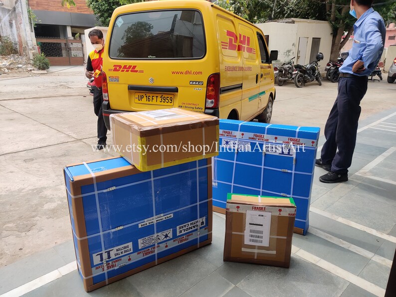 May include: Several packaged boxes, some wrapped in blue and brown cardboard, are stacked near a yellow DHL delivery van. The boxes are secured with tape and labeled with shipping information. The text "www.etsy.com/shop/IndianArtistArt" is visible.