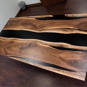 May include: A large wooden table with a black epoxy resin river running through the center. The table has a natural wood finish and is made from a single slab of wood.