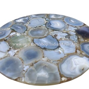 May include: A round, light blue agate table top with a natural, organic pattern. The table top is made of many pieces of agate that have been cut and polished to create a smooth surface. The agate is a light blue colour with white and brown accents. The table top is perfect for a modern or contemporary home.