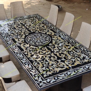 Puede incluir: Una mesa de comedor de mármol negro con una incrustación decorativa de nácar. La mesa está rodeada de ocho sillas con tapicería beige. La superficie de la mesa presenta un diseño floral con un medallón central.