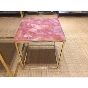 May include: A square pink and white marble top coffee table with a gold metal frame. The table is on a woven rug.