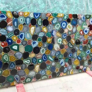 May include: A large rectangular slab composed of numerous polished agate stones. The stones are a mix of colors, including blue, green, yellow, black, and white, creating a mosaic-like pattern. The slab is likely for decorative or functional use.