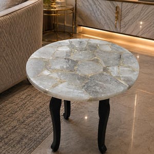 May include: A round side table featuring a tabletop constructed from irregular, translucent gray quartz stone fragments set in a smooth, polished surface. The table is supported by three curved, solid black legs that taper toward the floor.