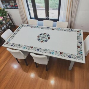 Op de afbeelding: Een rechthoekige witte eettafel met een bloemeninlegontwerp. De tafel heeft een centraal bloemenmedaillon en een rand van kleurrijke bloempatronen. Witte stoelen omringen de tafel, geplaatst op een houten vloer.