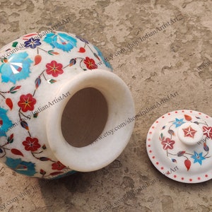 May include: White marble jar with a lid, decorated with intricate floral patterns in red, blue, and green. The jar is hand-painted with a floral design and has a lid with a star-shaped knob.
